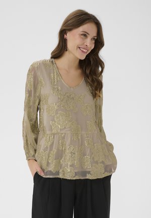 Woman smiling wearing a sheer beige blouse with gold floral patterns and black pants, hands in pockets, against a plain light background.