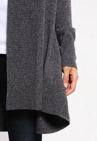 Cardigan ample gris en maille côtelée, avec un ourlet asymétrique et des manches longues, présentant une finition texturée.