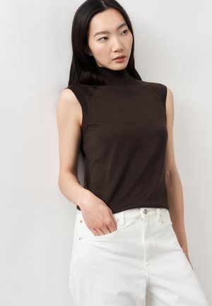 Ung kvinde med langt mørkt hår iført en ærmeløs sort rullekravetrøje og hvide bukser, stående med den ene hånd i lommen.