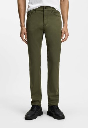 Mann trägt olivgrüne Slim-Fit-Hose und schwarze Sneakers, steht vor weißem Hintergrund.