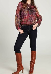 Blusa colorata con motivo floreale, maniche a palloncino e dettaglio con lacci al colletto, abbinata a jeans scuri e stivali alti in pelle marrone.