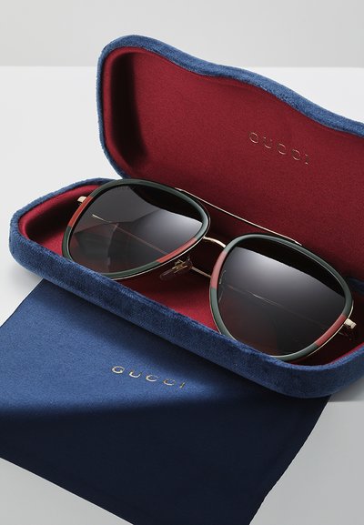 Gucci solbriller i et blått fløyelsetui med rødt interiør. Har gulltilbehør, store mørke linser og en grønn og rød ramme.