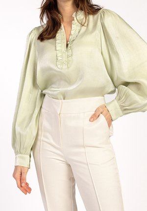Femme portant un chemisier en satin vert clair avec patte de boutonnage volantée et manches bouffantes, associé à un pantalon crème taille haute.
