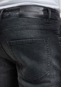 Nærbilde av baklommen og midjebåndet på svarte denimjeans med svart lærmerke preget med "ZY PREMIUM", brukt med en hvit topp.