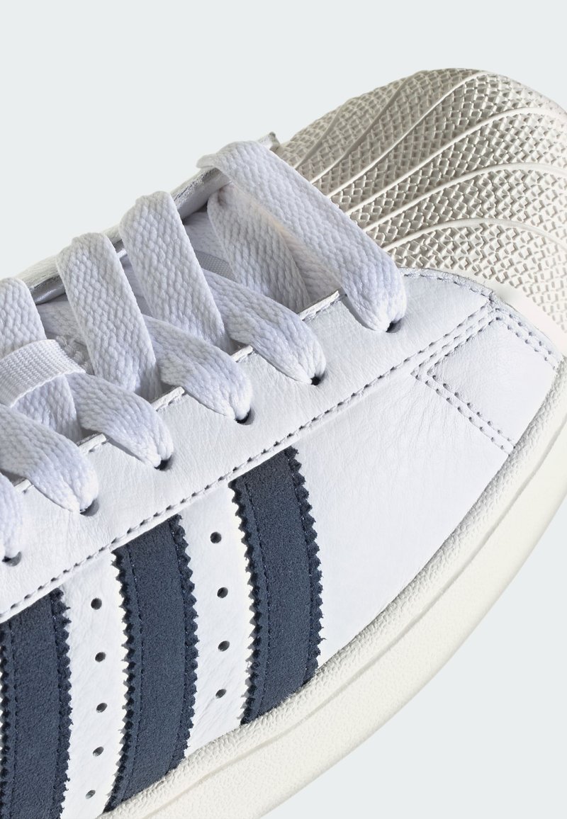 White sneaker with thick textured rubber toe cap, white laces, and three black vertical stripes on leather upper side.