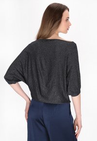 Černý, třpytivý volný top s křidélkovými rukávy. Hladký materiál, off-shoulder design, spárovaný s modrými volnými kalhotami.