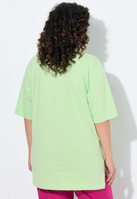 T-shirt oversize verde menta con maniche corte. Tessuto morbido, orlo arrotondato e spacchetti laterali, indossato con pantaloni rosa. La vista da dietro mostra capelli ricci.