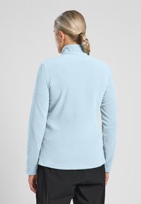 Błękitny polarowy sweter z wysokim kołnierzem i długimi rękawami, charakteryzujący się miękką fakturą i zaokrąglonym dołem na wysokości talii, noszony na ciemnych spodniach.