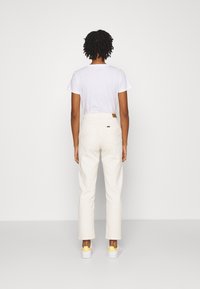 Ljusbeige byxor med hög midja och rak skärning, med bakfickor och en tanfärgad läderetikett på midjebandet, kombinerade med vita skor.