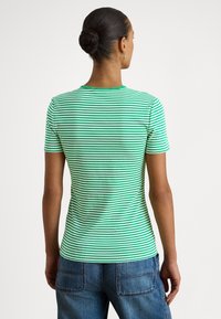 Žena s vlasmi zaviazanými do drdola, oblečená v zeleno-biele pruhovanej krátkosleevej košeli a modrých džínsoch, viditeľná zozadu na jednoduchom pozadí.