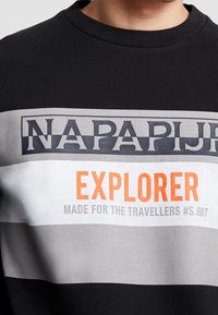 Sudadera negra con rayas horizontales en gris y blanco, con "NAPAPIJRI" en negrita y "EXPLORER" en naranja. Tejido de algodón, cuello redondo.