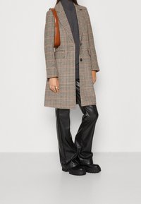 Manteau long à carreaux en beige et crème avec un col à revers, porté sur un col roulé foncé, associé à un pantalon noir en similicuir et des chaussures épaisses.