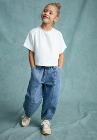 Vit kortärmad t-shirt som är croppad, kombinerad med löst sittande blå denim-joggers och beige sneakers med vita snören och detaljer.