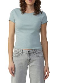Frau in einem hellblauen, kurzärmeligen Ripp-Top und hoch geschnittenen, hellgrauen Jeans vor einem weißen Hintergrund.