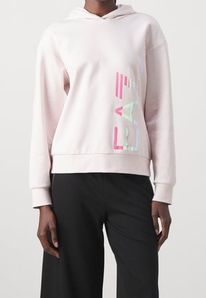 Persoon die een lichtroze capuchontrui draagt met meerkleurige verticale letters aan de rechterkant en zwarte wijde broekspijpen.