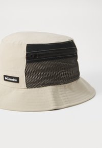 Beige bucket hat featuring a zippered black mesh accent and logo patch. Material is soft fabric with a slightly glossy finish.