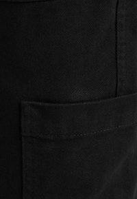 Svart denimtyg med en texturerad yta, med en synlig ficka och subtila sömnadsdetaljer. Fokusera på materialet och konstruktionen.