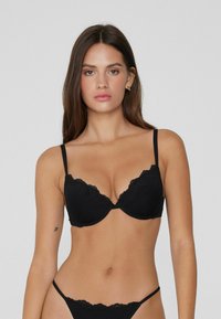Žena stojící proti jednoduchému pozadí, oblečená v černé podprsence s krajkovým lemem a odpovídajícím spodním prádlem, hledící vpřed s neutrálním výrazem.
