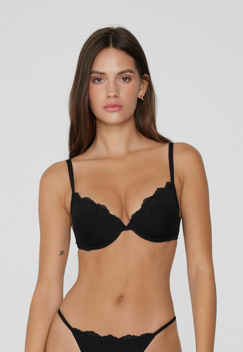 Žena stojící proti jednoduchému pozadí, oblečená v černé podprsence s krajkovým lemem a odpovídajícím spodním prádlem, hledící vpřed s neutrálním výrazem.