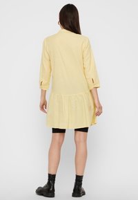 Gelbes, midilanges Kleid mit dreiviertel Ärmeln, mehrstufigem Rock und seitlichen Schlitzen, gefertigt aus leichtem Stoff. Über schwarzen Leggings getragen.