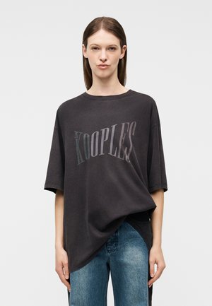 Kvinde iført oversized sort T-shirt med "The Kooples"-logo og blå jeans, stående mod en ensfarvet lys baggrund.