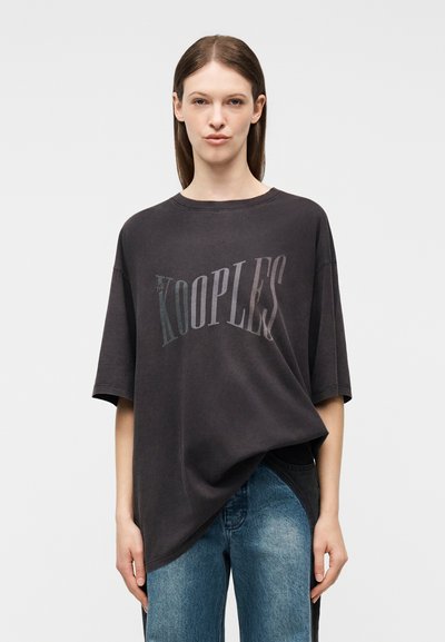 Γυναίκα που φοράει υπερμεγέθη μαύρο T-shirt με το λογότυπο "The Kooples" και μπλε τζιν, στέκεται μπροστά σε ένα απλό φωτεινό φόντο.
