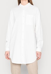 Chemise blanche à manches longues en tissu texturé, avec un col pointu, une fermeture à boutons et une poche poitrine unique.
