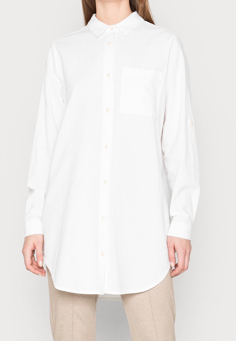 Chemise blanche à manches longues en tissu texturé, avec un col pointu, une fermeture à boutons et une poche poitrine unique.