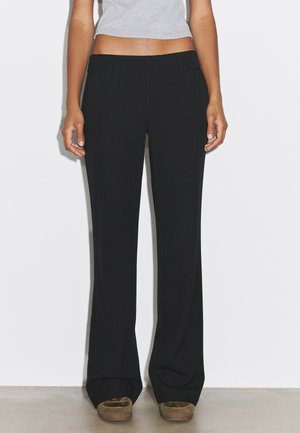 Femme portant un pantalon noir à jambes larges, un top court gris et des mocassins marron, debout devant un fond blanc uni.