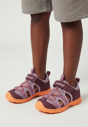 Dziecko nosi fioletowo-pomarańczowe sandały Hummel z odkrytymi palcami i beżowe krótkie spodenki, stoi na białej powierzchni.
