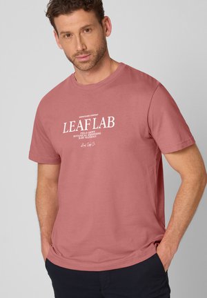 Man with short curly hair wearing a dusty rose T-shirt with "LEAF LAB" white text, hands in pockets, neutral gray background.