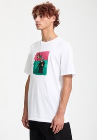 Hvid bomulds T-shirt med et farverigt grafisk print med abstrakte figurer og en solid sort kant. Casual pasform og korte ærmer.