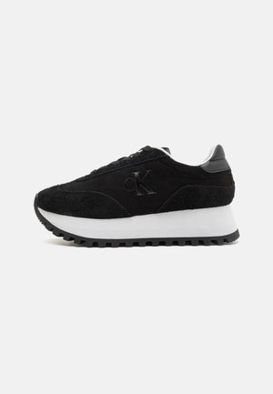 Zapatilla de ante negro con suela de goma blanca y negra, que presenta un upper texturizado, punta redonda y un logo bordado en el costado.