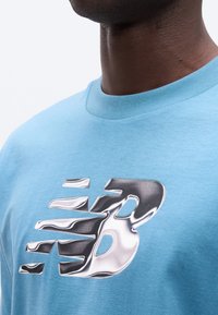 Κοντινό πλάνο ατόμου που φοράει ανοιχτό μπλε πουκάμισο με μεγάλο αντανακλαστικό ασημί λογότυπο "NB" στο στήθος, με απλό λευκό φόντο.