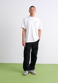 Homme debout sur un sol vert, portant un T-shirt blanc surdimensionné, un pantalon cargo noir et des baskets argentées, regardant droit devant lui avec une expression neutre.