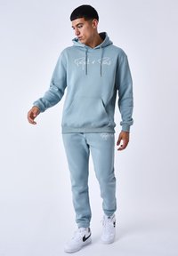 Felpa leggera blu con pantaloni abbinati in tessuto morbido, dotata di tasca frontale, cordini e dettagli del logo in bianco. Le sneakers sono bianche con accenti neri.