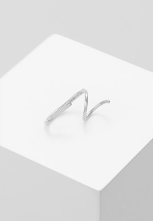 Einfacher silberner gedrehter Ring mit offenem Spiralmuster, platziert auf einem weißen würfelförmigen Sockel vor hellem grauem Hintergrund.