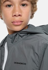 Grå lättviktsjacka med huva, med dragkedja och svart "DCSHOECO"-logotyp framtill. Slät textur och elegant design.