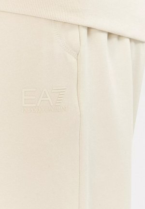 Pantaloni della tuta beige realizzati in tessuto morbido, con vita a coste, tasche laterali e logo EA7 in rilievo sotto la linea della vita.