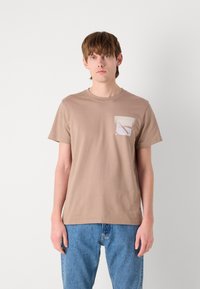 T-shirt en coton beige avec un col rond et des manches courtes, présentant une poche contrastante avec un design géométrique. Porté avec un jean bleu.