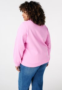 Kruidige vrouw met krullend haar, gekleed in een roze sweatshirt met geborduurde harten op de mouwen en blauwe jeans, zijwaarts kijkend op een witte achtergrond.
