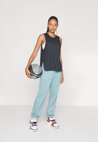Top senza maniche nero con fessure laterali, pantaloni sportivi blu chiaro e sneakers multicolori, mentre tiene un pallone da basket di fronte a uno sfondo semplice.