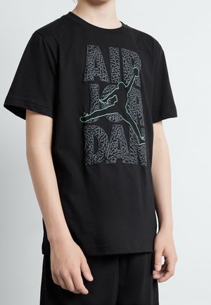 Barn iført sort T-shirt med stort gråt tekstureret "AIR JORDAN" logo og en grøn optegnet silhuet af en basketballspiller.