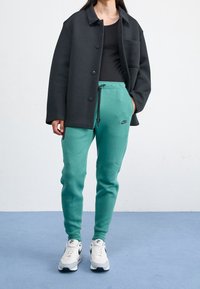 Černá oversize košile s knoflíky přes černé tílko, v kombinaci s tyrkysovými tepláky s tkaničkou a logem Nike; bílé tenisky.