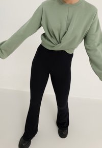 Persona vestida con un sudadera verde claro retorcida a la altura de la cintura, pantalones de campana negros con relieve y zapatos gruesos negros sobre un fondo uniforme.