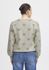 Pull beige avec motifs de cœurs verts, col côtelé et manches bouffantes. Associé à un jean en denim bleu clair.