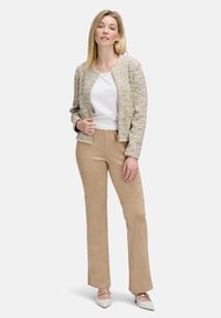Kobieta wearing textured beige jacket, white top, tan suede pants, and white heels standing against a plain light background.