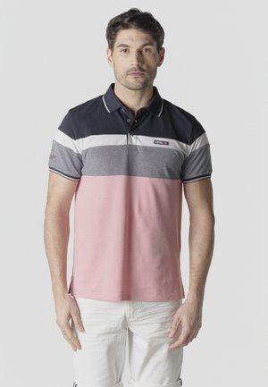 Hombre de pie mirando hacia adelante, vistiendo una camisa tipo polo de bloques de color en azul marino, blanco, gris y rosa, con pantalones blancos.