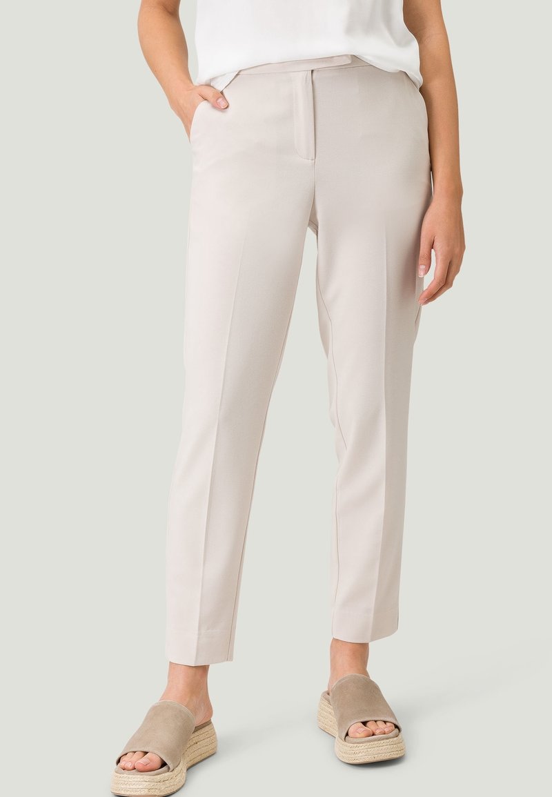 Pantalons beiges fuselés à coupe ajustée, avec poches latérales et devant épuré. Texture lisse avec une apparence légère, associés à des chaussures ouvertes beiges.