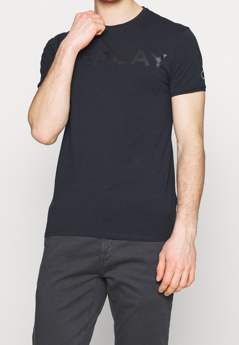 Mörkblå bomullströja med rund halsringning, med en texturerad, mörkgrå logotyp över bröstet och en liten logotyp på ärmen.